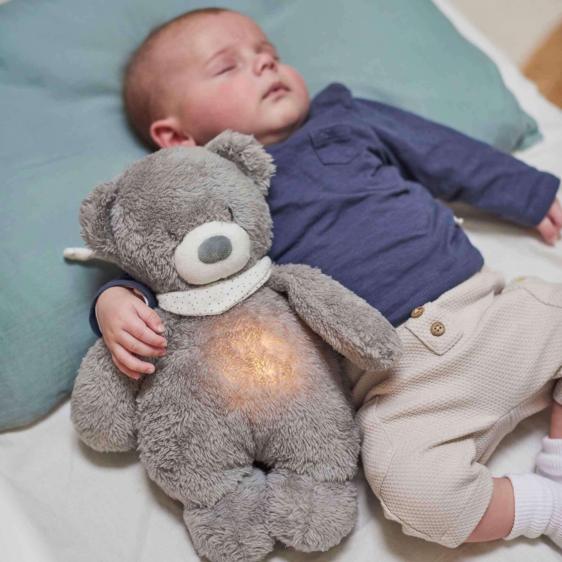 Baby sleeping peacefully while cuddling a Nattou grey Sleepy Cuddly bear nightlight, which emits a soft warm glow from its belly. Ideal for bedtime comfort.