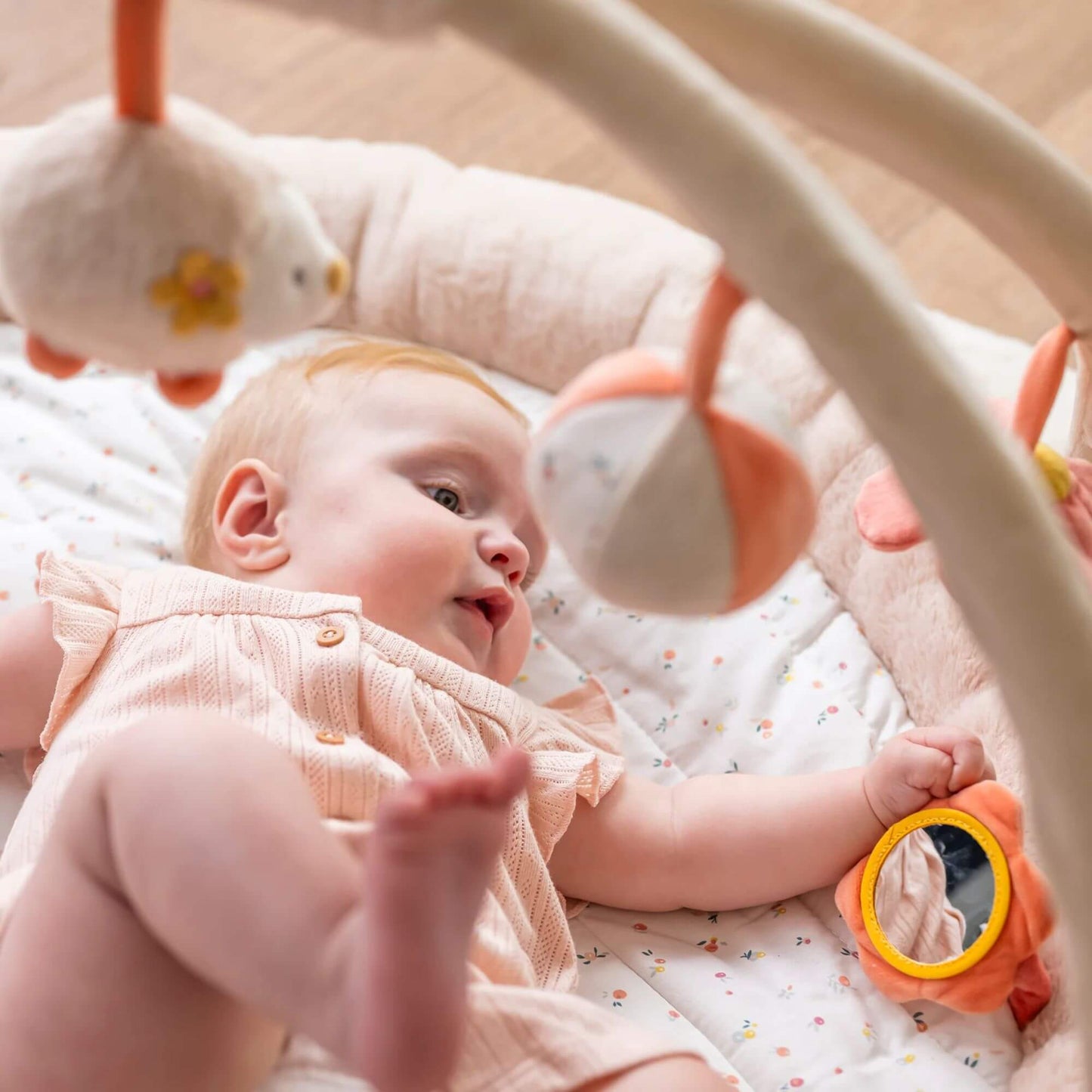 Baby lying on the Nattou Mila, Lana & Zoe playmat, reaching for hanging plush toys and a soft ball, with a flower-shaped mirror toy nearby for sensory play.