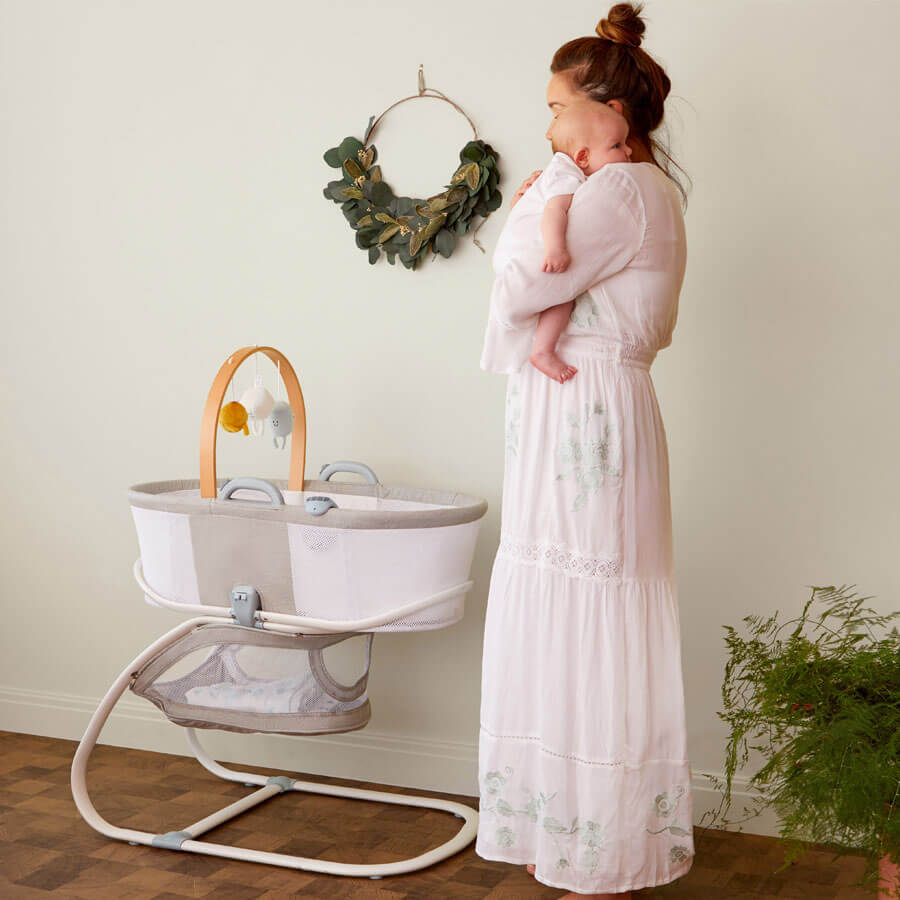 Mother holding a baby next to the Purflo breathable crib, featuring a modern beige and grey design, mesh sides, wooden mobile arch with plush toys, and storage basket.