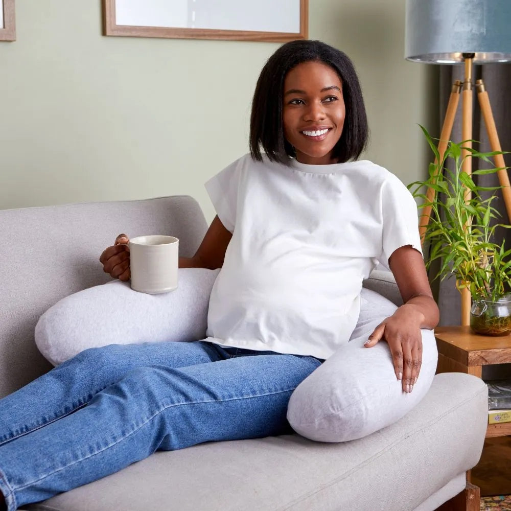 Woman relaxing on a sofa with a cup of tea, using the Purflo Breathe Pregnancy Pillow in minimal grey for ergonomic support and comfort.