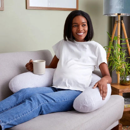 Woman relaxing on a sofa with a cup of tea, using the Purflo Breathe Pregnancy Pillow in minimal grey for ergonomic support and comfort.