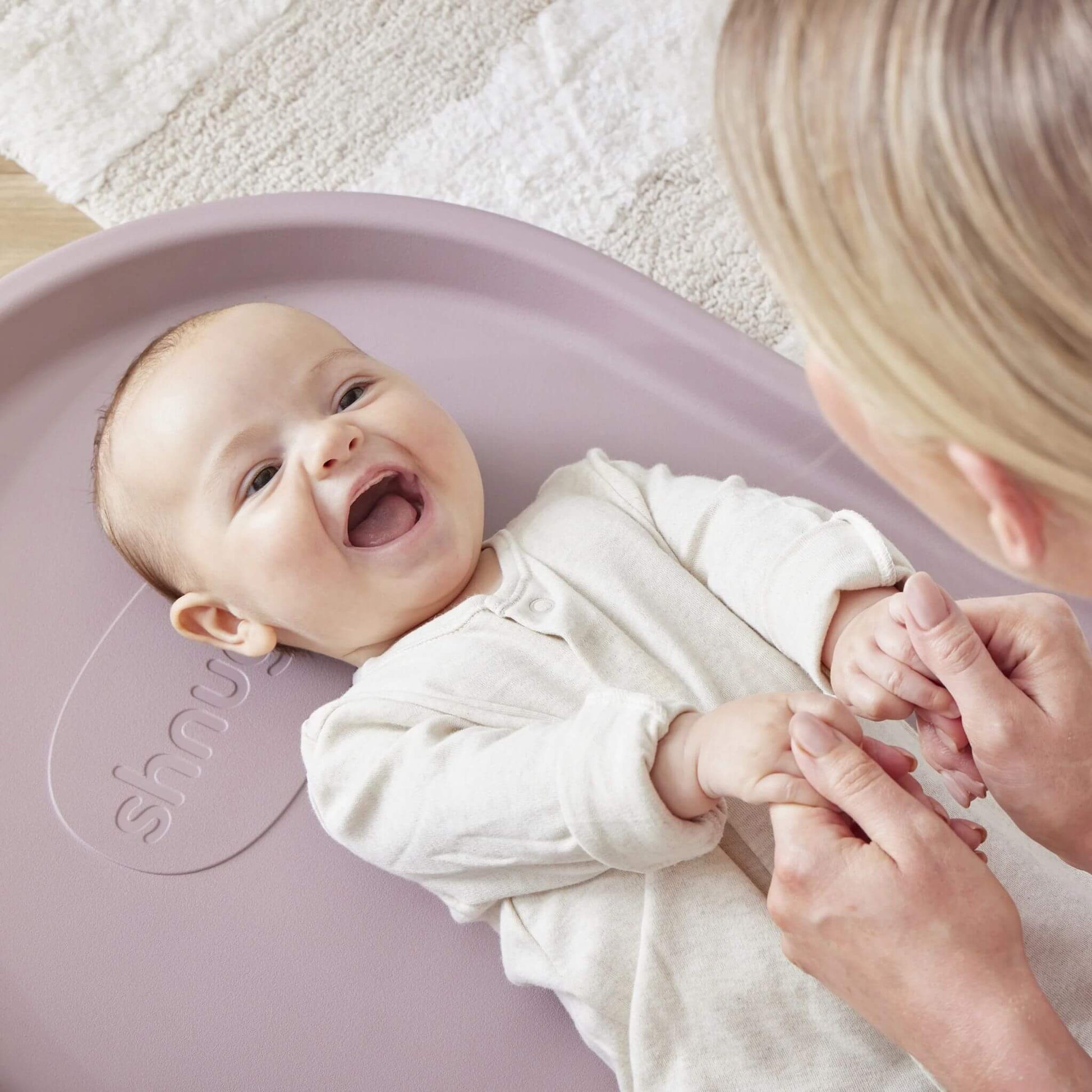 Smiling baby lying on blossom Shnuggle Squishy Changing Mat wearing white sleepsuit, mother's hands visible during nappy change moment.