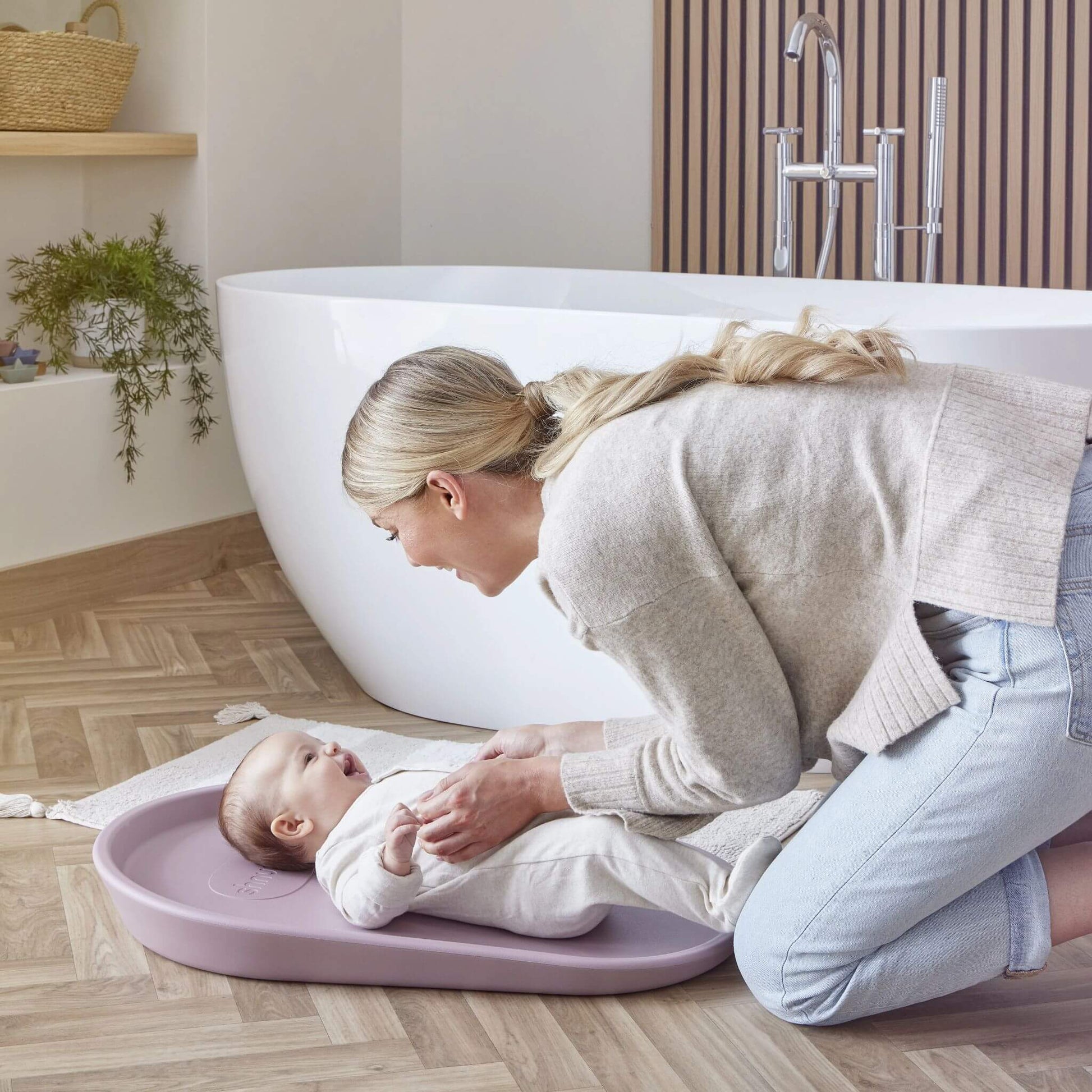 Shnuggle Squishy Change Mat in modern bathroom setting with freestanding bath, mother changing baby's nappy on soft, waterproof surface.