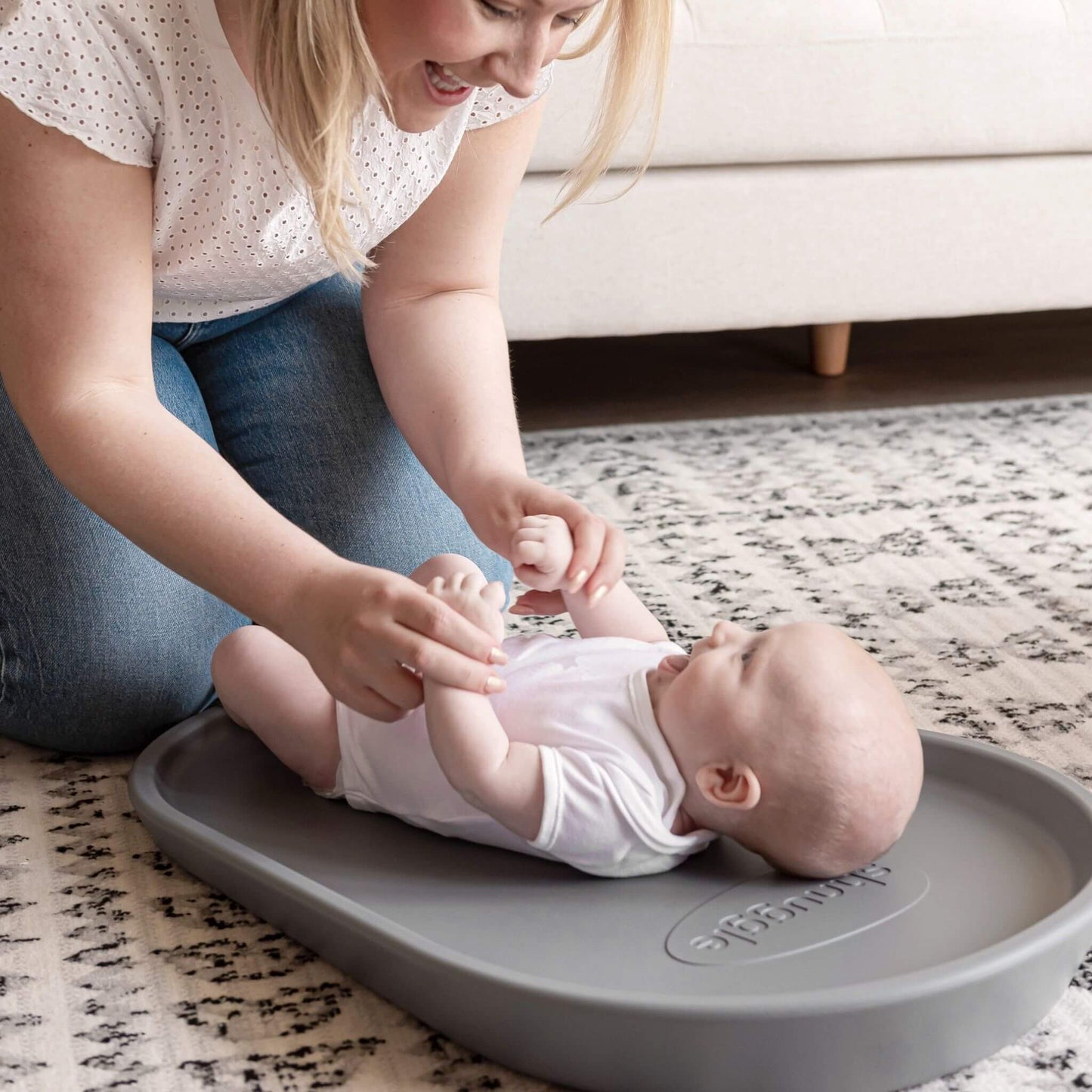 Mother smiling and holding baby's hands while the baby lies on the Shnuggle Squishy Changing Mat in grey, creating a bonding moment.