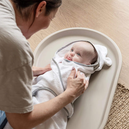 Mother smiling at baby wrapped in hooded towel on Shnuggle Squishy Changing Mat in taupe, providing comfort during nappy change.