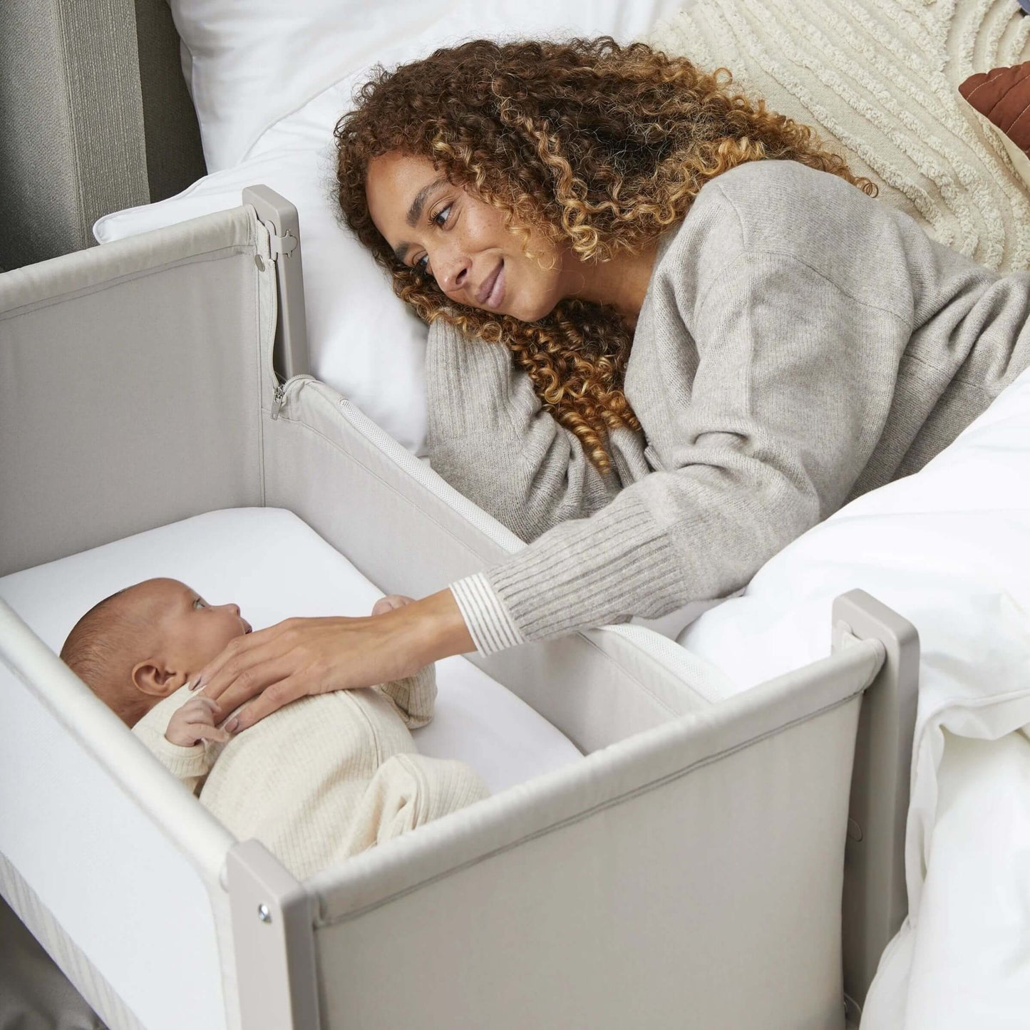 Mother reaches out to baby in Shnuggle Air Lite Bedside Crib, allowing close contact and safe co-sleeping with breathable mesh panel.