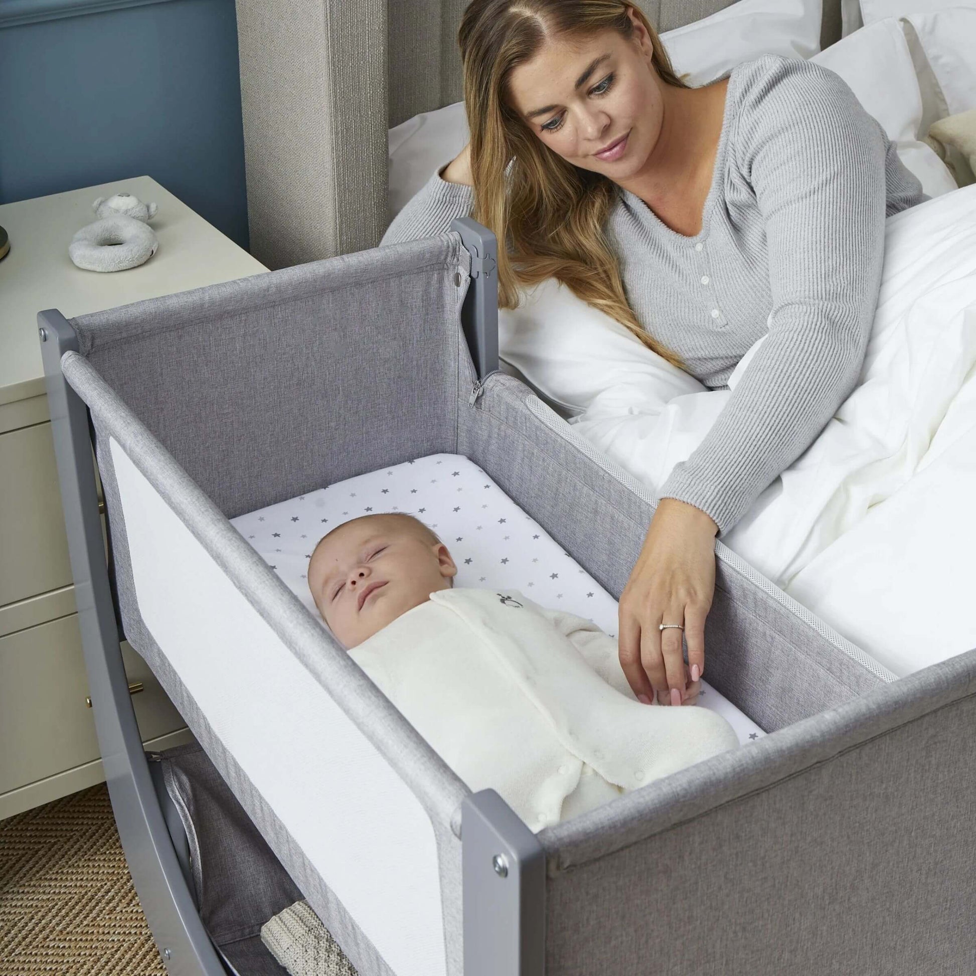 Mother gently touches sleeping baby in Shnuggle Air Lite Bedside Crib, promoting close and safe co-sleeping with breathable mesh sides.