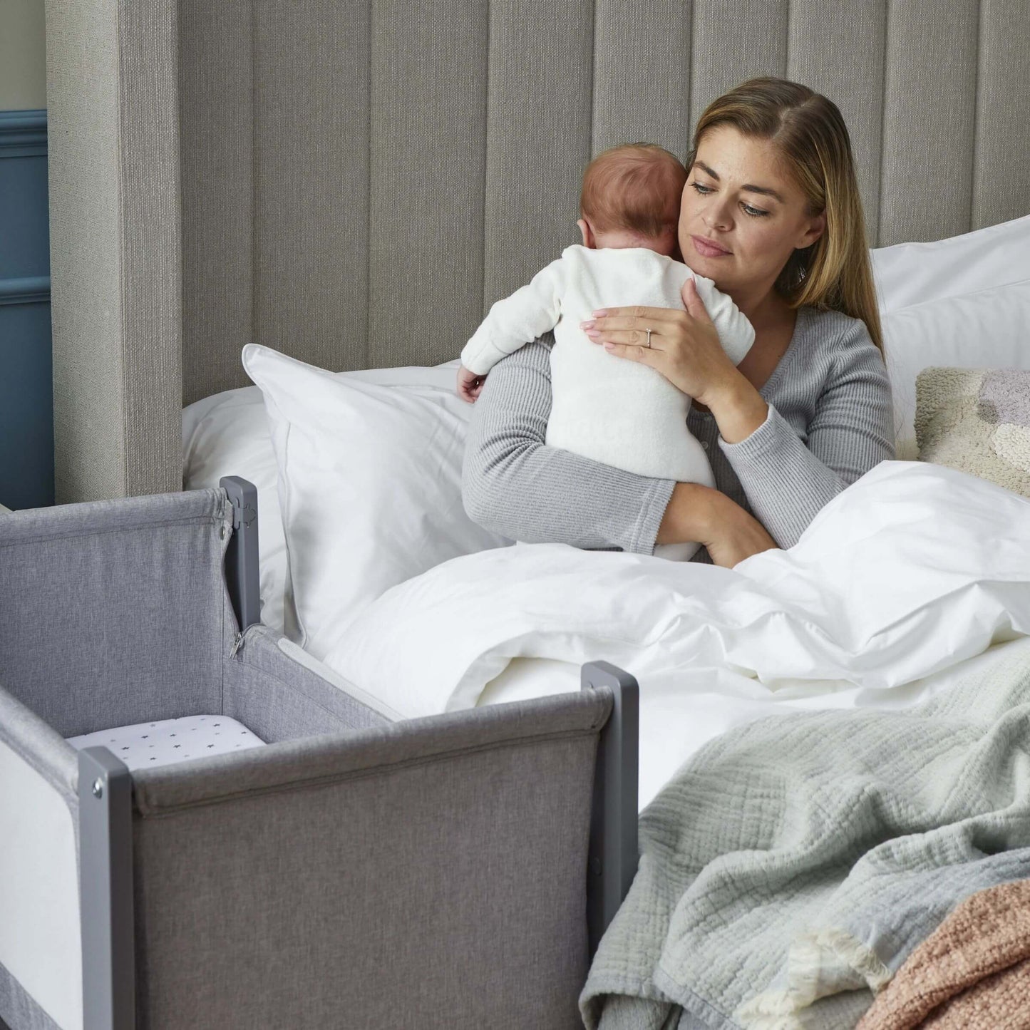 Mother sitting up in bed, holding baby close beside Shnuggle Air Lite Bedside Crib, emphasising easy access and bonding.