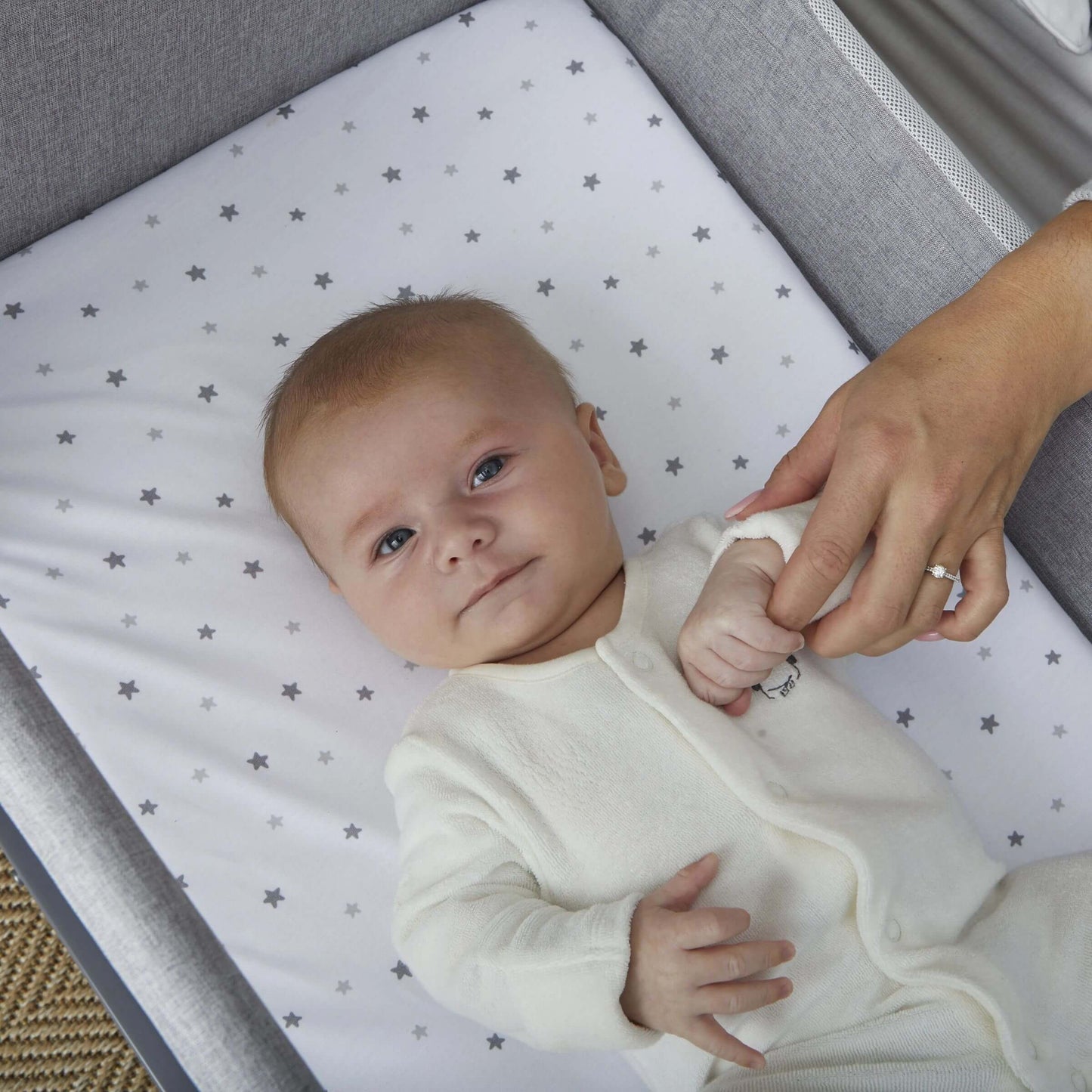 Baby lying in Shnuggle Air Lite Bedside Crib, holding mother's hand, promoting comfort and connection.