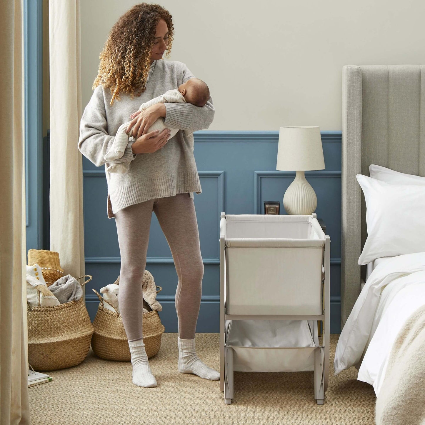 Mother holding baby beside Shnuggle Air Lite Bedside Crib in bedoom.