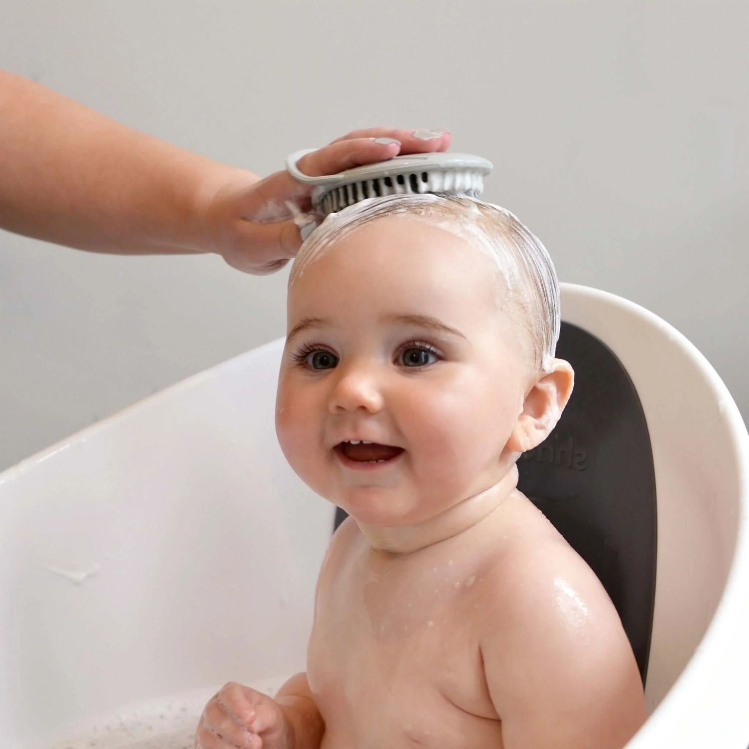 Happy baby sitting in a bath while a mother gently uses the Shnuggle baby bath brush to lather the baby’s scalp, creating a calming experience.