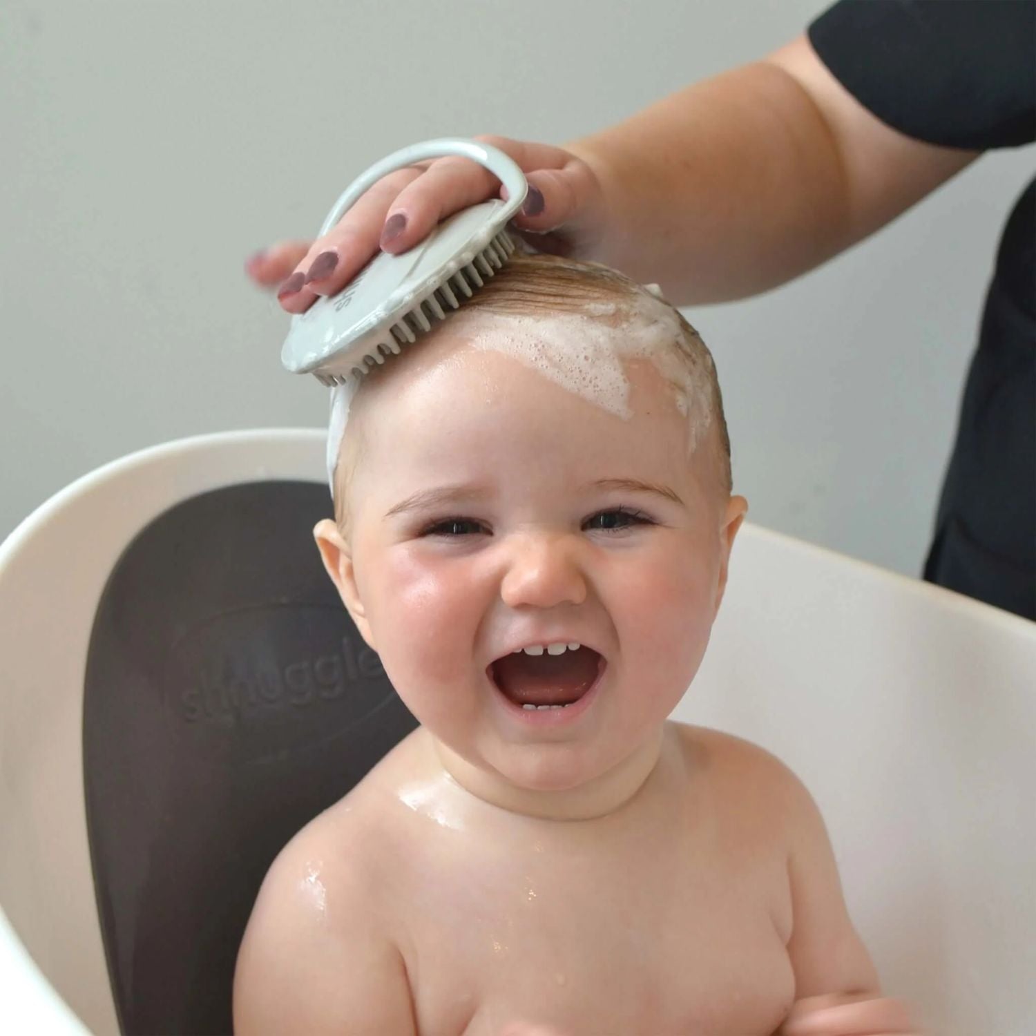 Smiling baby in a bath while a mother gently uses the Shnuggle baby bath brush to lather and massage the baby’s scalp.