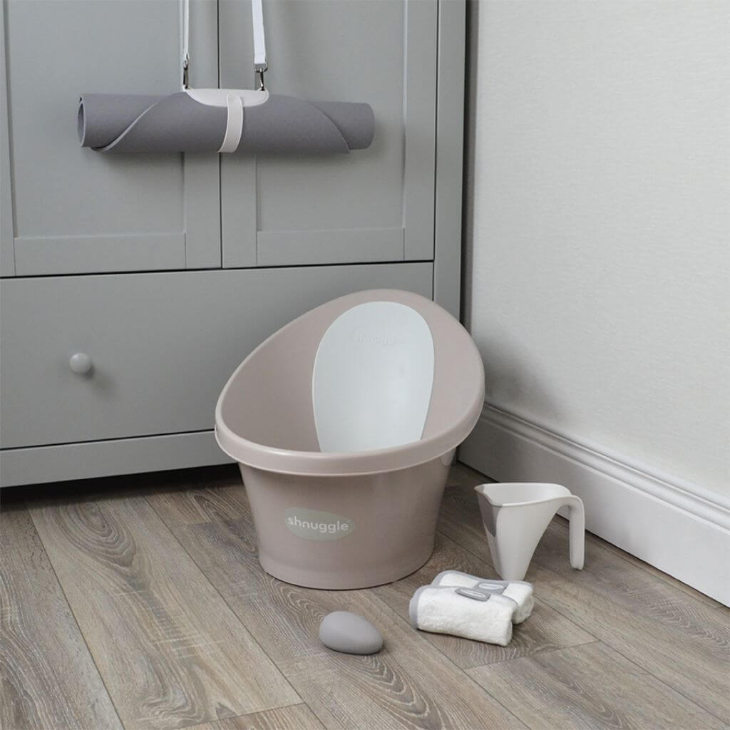 Shnuggle baby bath in taupe with Bum Bump support, placed in a nursery beside soft towel, water jug, and baby essentials for bath time.