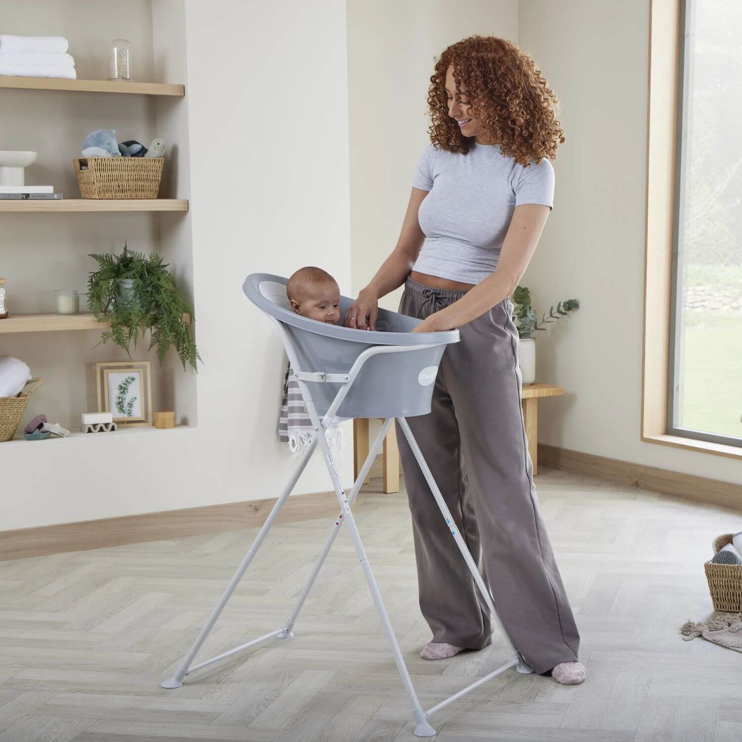 Smiling mother bathes a baby in a grey Shnuggle baby bath on a tall stand, in a bright and cosy bathroom setting.