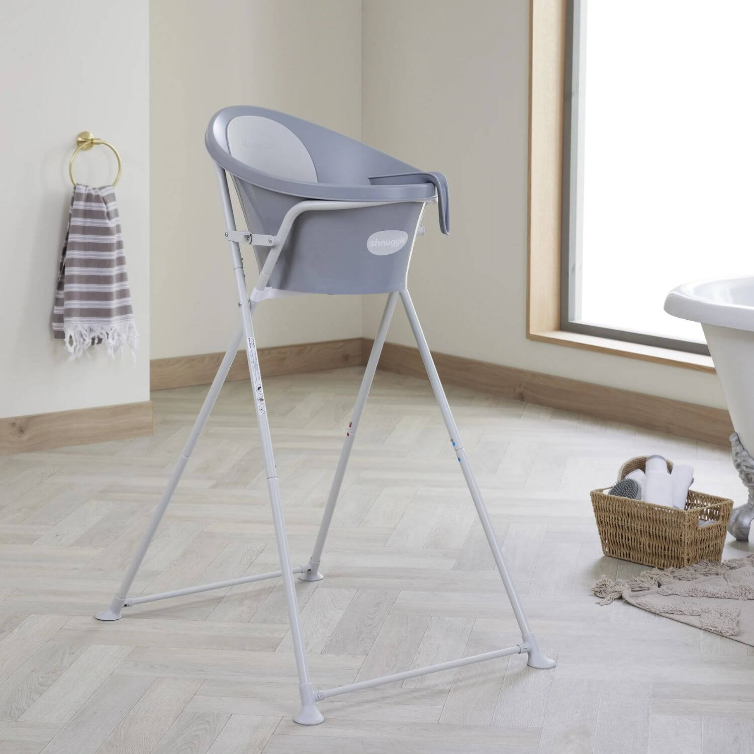 Grey Shnuggle baby bath securely placed on a tall stand in a cosy bathroom setting, with a towel and basket nearby.