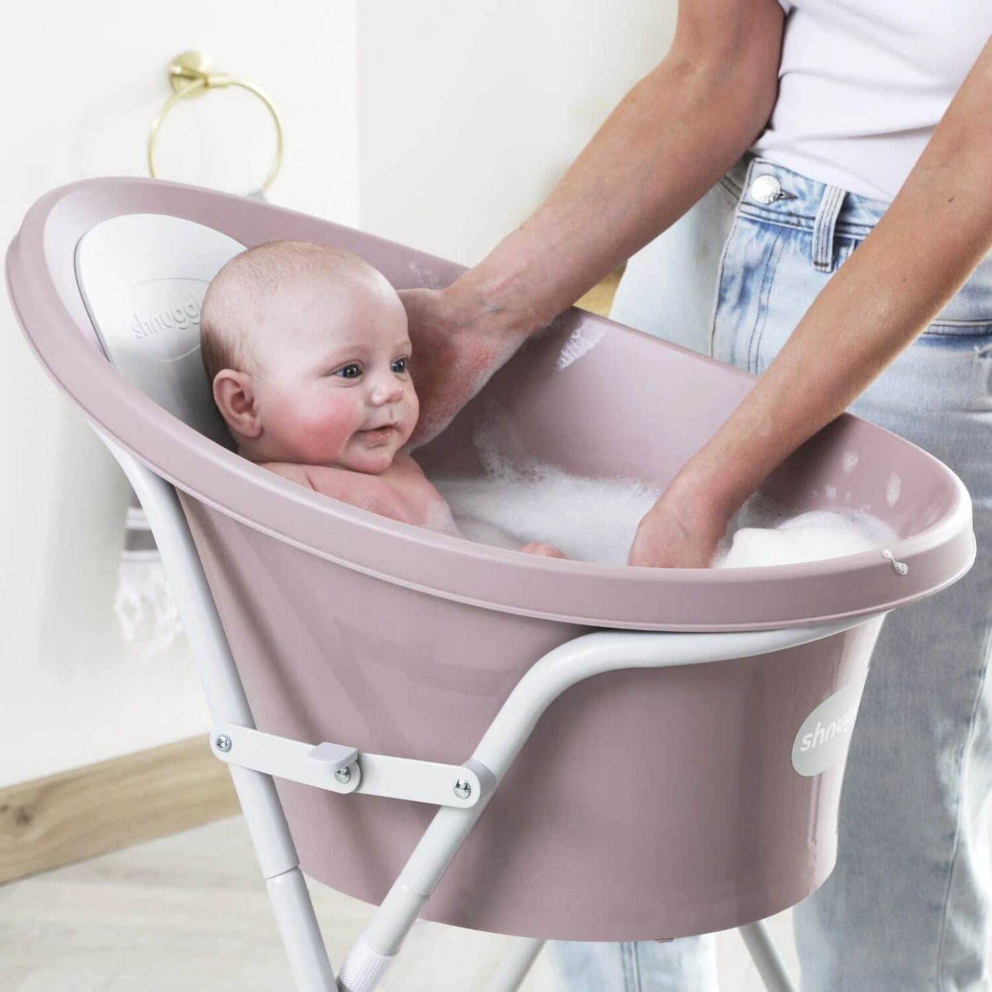 Smiling baby enjoys a bath in a pink Shnuggle baby bath placed on a tall stand, with a caregiver’s hands gently supporting them.