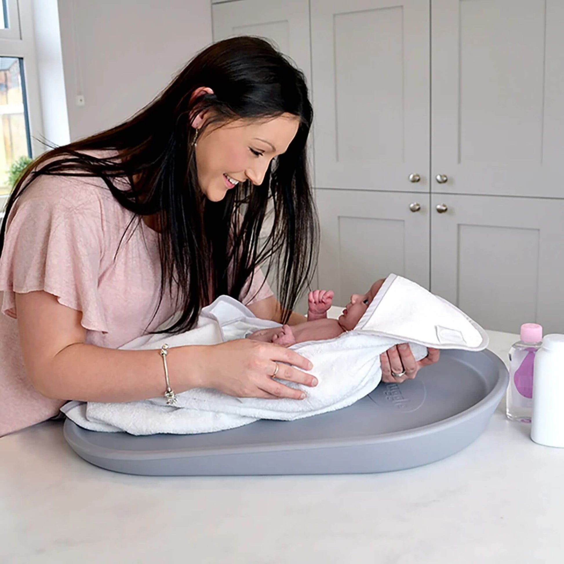 Mother smiling at her newborn wrapped in a towel on the Shnuggle Squishy Changing Mat in grey, creating a warm bonding moment.