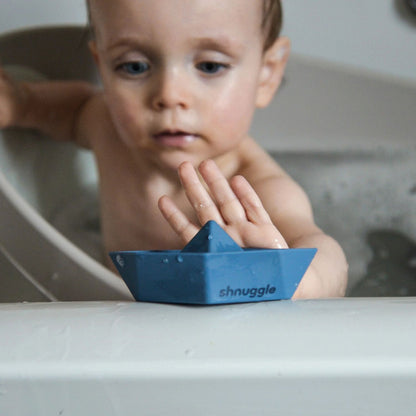 Child reaching out to touch a blue Shnuggle Stack ‘n’ Sail Bath Boat Toy, engaging in playful bath time fun.