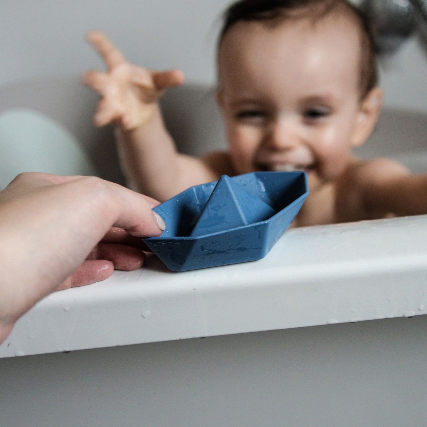 Happy child in the bath reaching for a blue Shnuggle Stack ‘n’ Sail Bath Boat Toy, engaging in playful interaction with a caregiver.