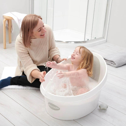 Laughing toddler splashes in a Shnuggle bath while a smiling caregiver sits nearby on the bathroom floor. A towel rests on a stool in the background.