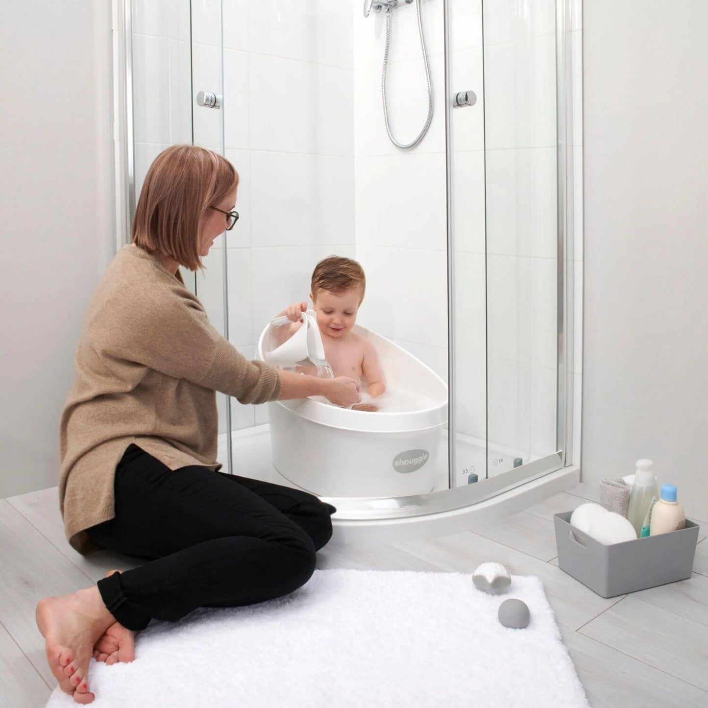 Toddler sits in a Shnuggle bath, pouring water from a scoop, while a caregiver kneels beside the bath. Toiletries are in a box nearby.