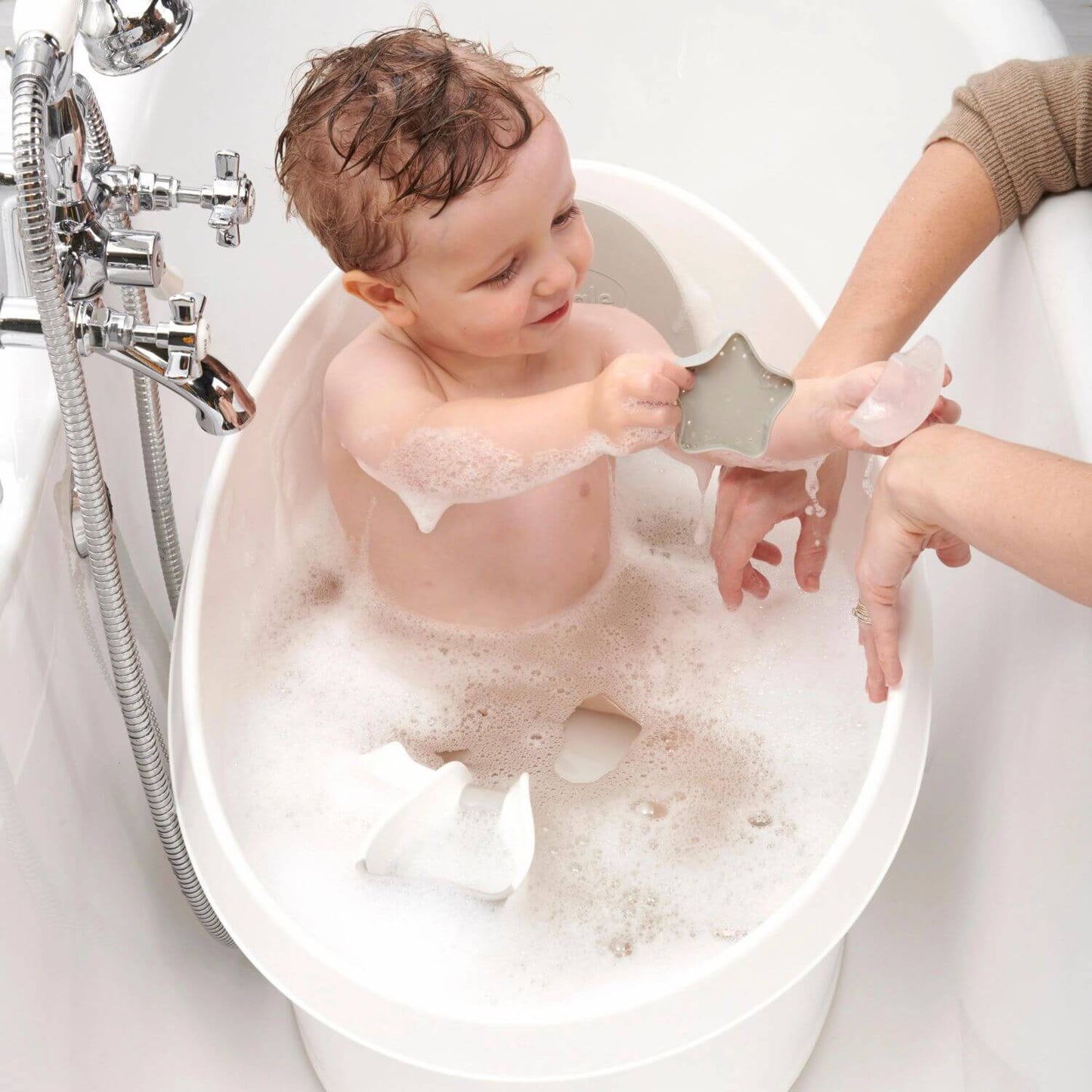 Smiling toddler sitting in a Shnuggle bath filled with bubbles, playing with a star-shaped toy while a caregiver's hand reaches in.