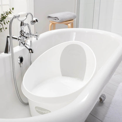 Shnuggle toddler bath placed inside a large bathtub, filled with water and bubbles. Chrome taps are visible, along with a folded grey towel on a small stool in the background.