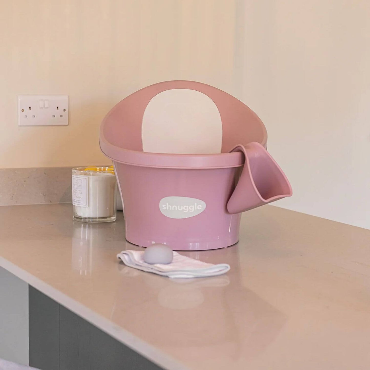 Pink Shnuggle baby bath with matching Washy bath jug attached, set on a countertop with a candle, cloth, and bath sponge nearby.