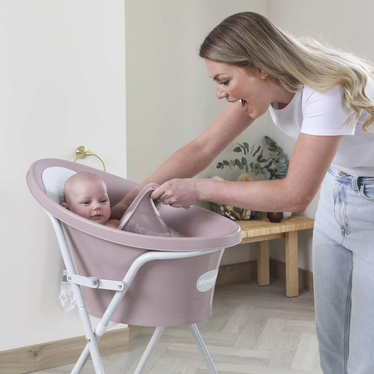 Smiling mother uses a pink Shnuggle Washy bath jug to rinse a baby in a matching Shnuggle bath on a stand, in a cosy bathroom setting.