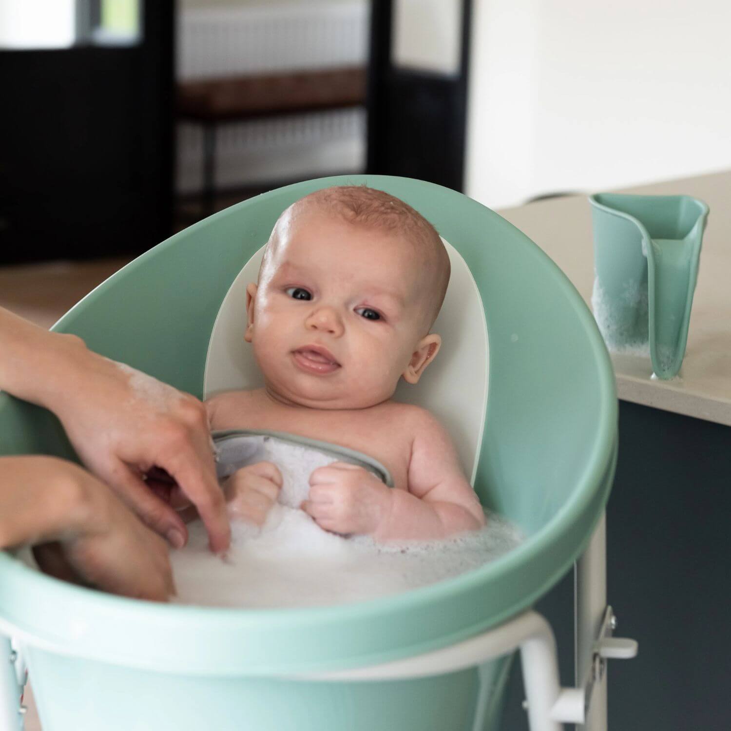 Baby in a eucalyptus Shnuggle bath with bubbles, being bathed by mother. Matching Shnuggle Washy bath jug is placed nearby on the counter.