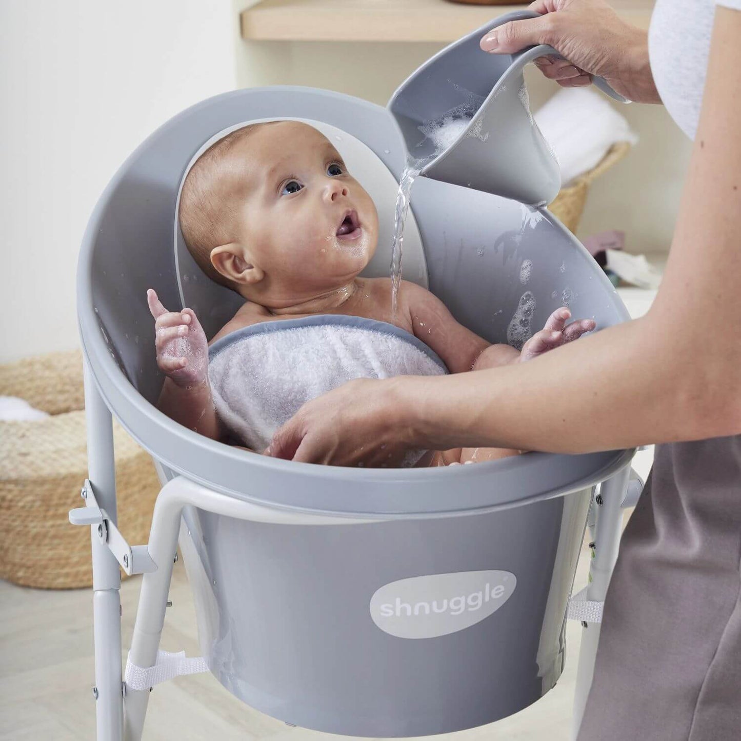 Caregiver uses a pebble grey Shnuggle Washy bath jug to rinse a baby in a matching Shnuggle bath on a stand, in a cosy bathroom setting.