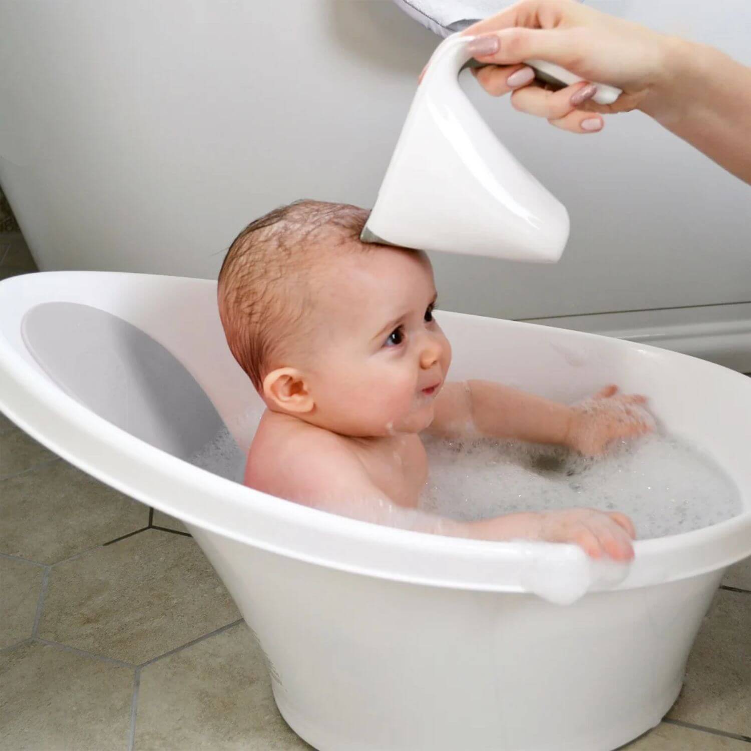 Mother gently rinses a smiling baby's hair with a white Shnuggle Washy bath jug, as the baby sits in a white Shnuggle bath filled with bubbles.