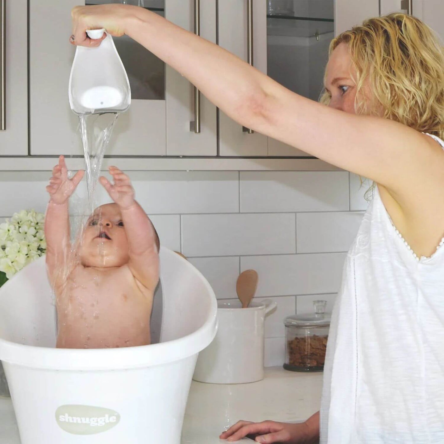 Mother pours water over a playful baby using a white Shnuggle Washy bath jug, as the baby reaches up in a white Shnuggle bath.