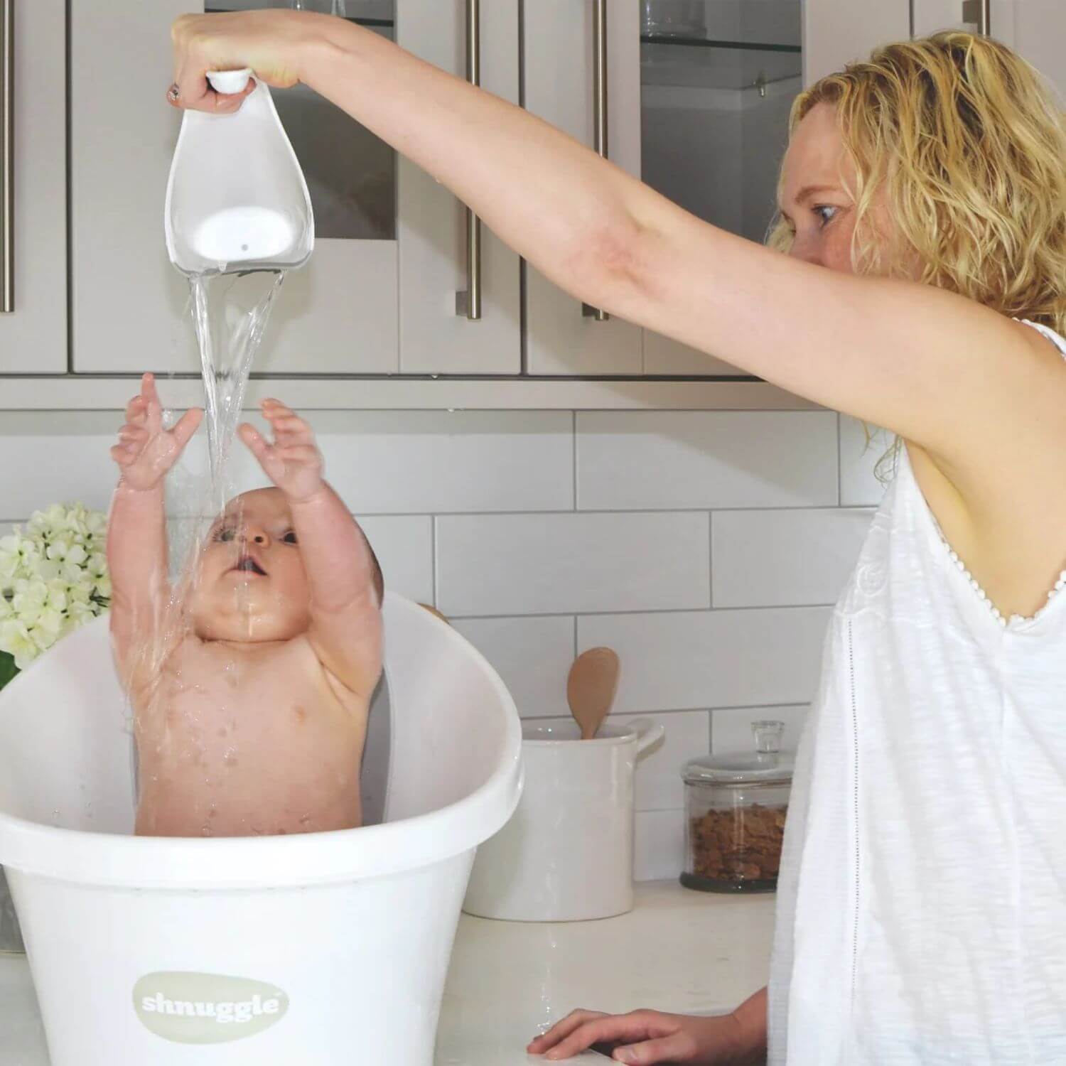 Mother pours water over a playful baby using a white Shnuggle Washy bath jug, as the baby reaches up in a white Shnuggle bath.