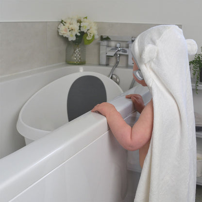 Baby wearing a Shnuggle wearable bath towel with hood and cute ears, standing by the bathtub ready for bath time.