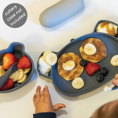 Toddler using grey TUM TUM bear-shaped plate and snack pot with pancakes, fruit, and yoghurt,  perfect for self-feeding.