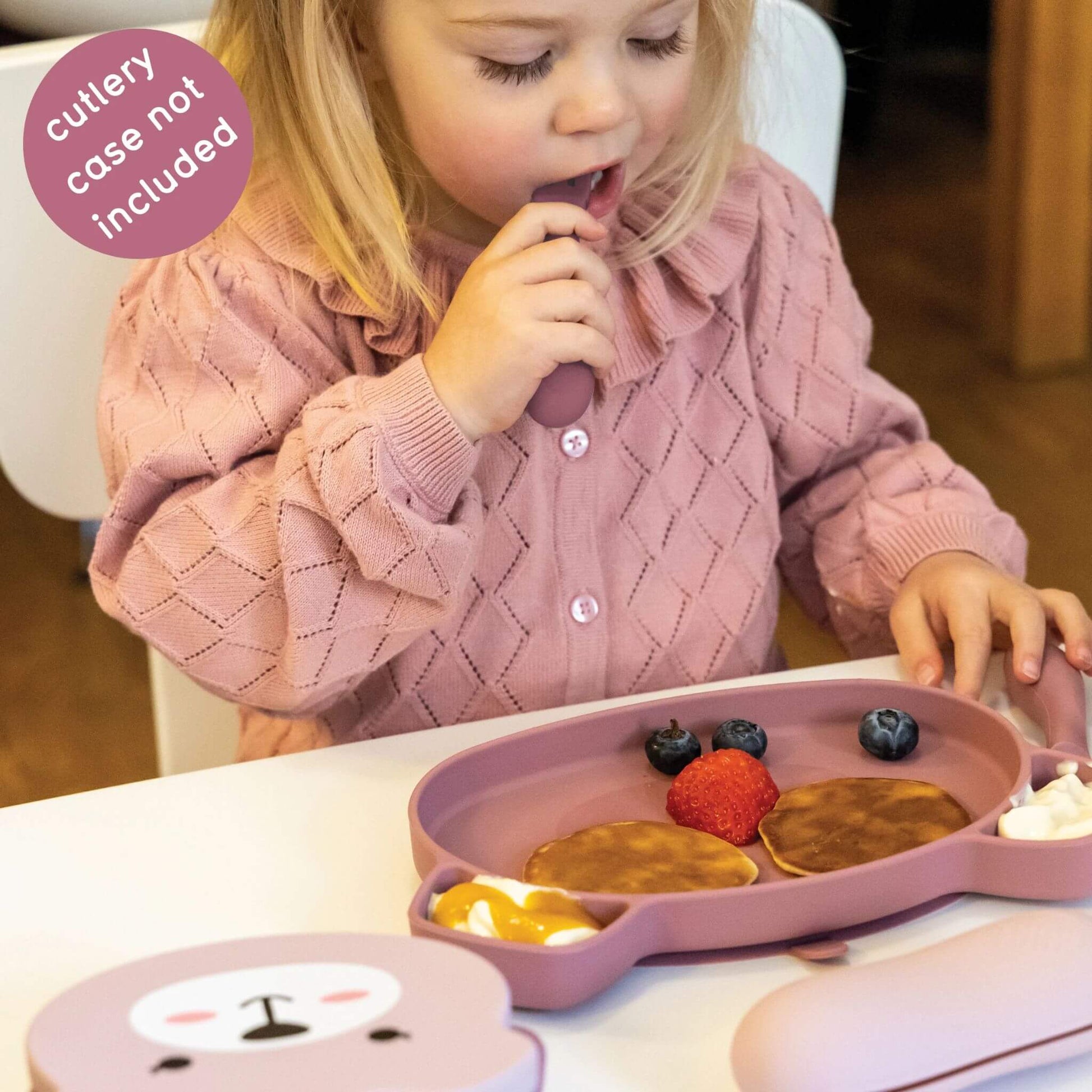 Toddler using pink TUM TUM silicone cutlery and plate with pancakes, fruit, and yoghurt, wearing a pink jumper, perfect for self-feeding.