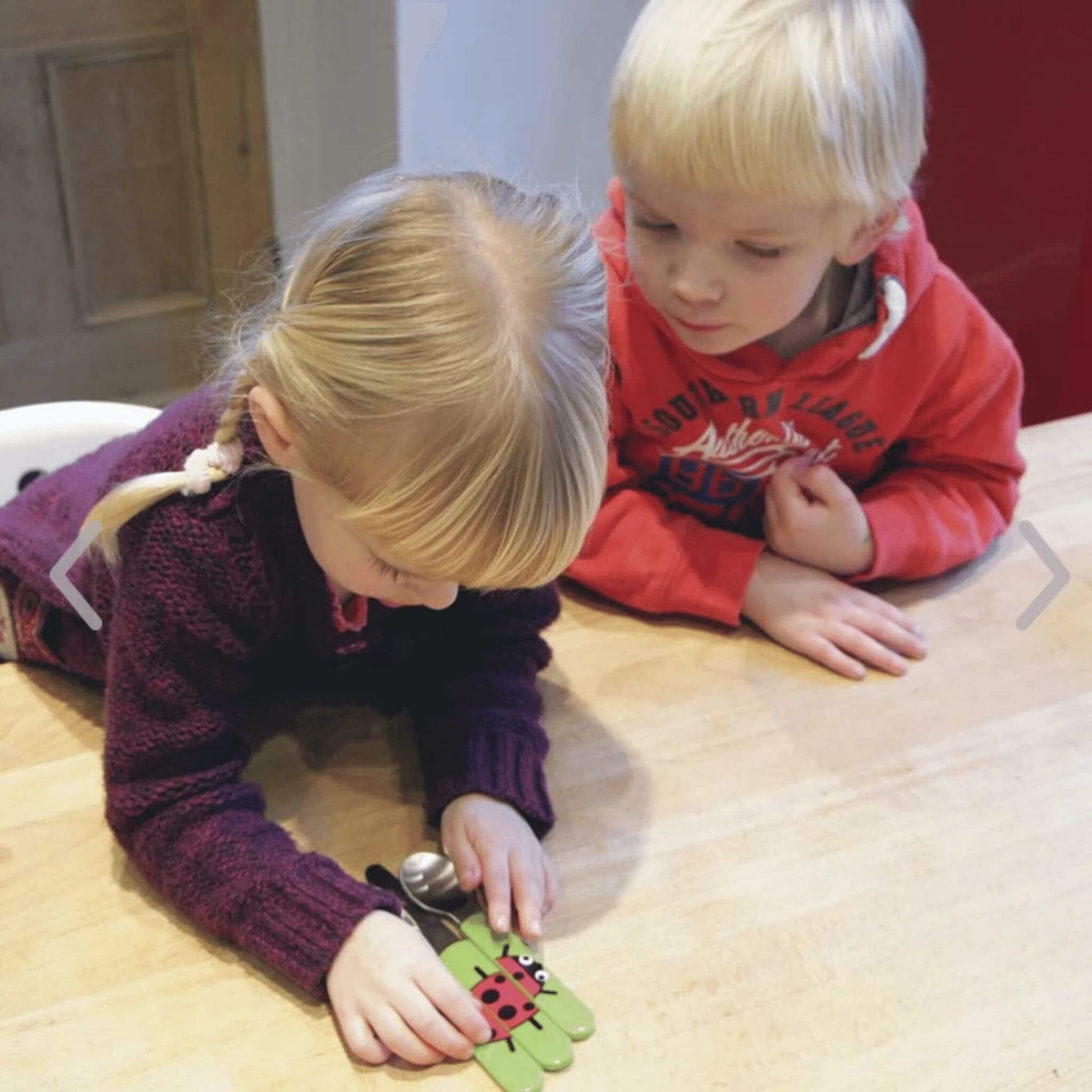 Two children examining the TUM TUM Easy Scoop Cutlery Set, featuring bright green handles with a ladybird design. Ideal for encouraging self-feeding.