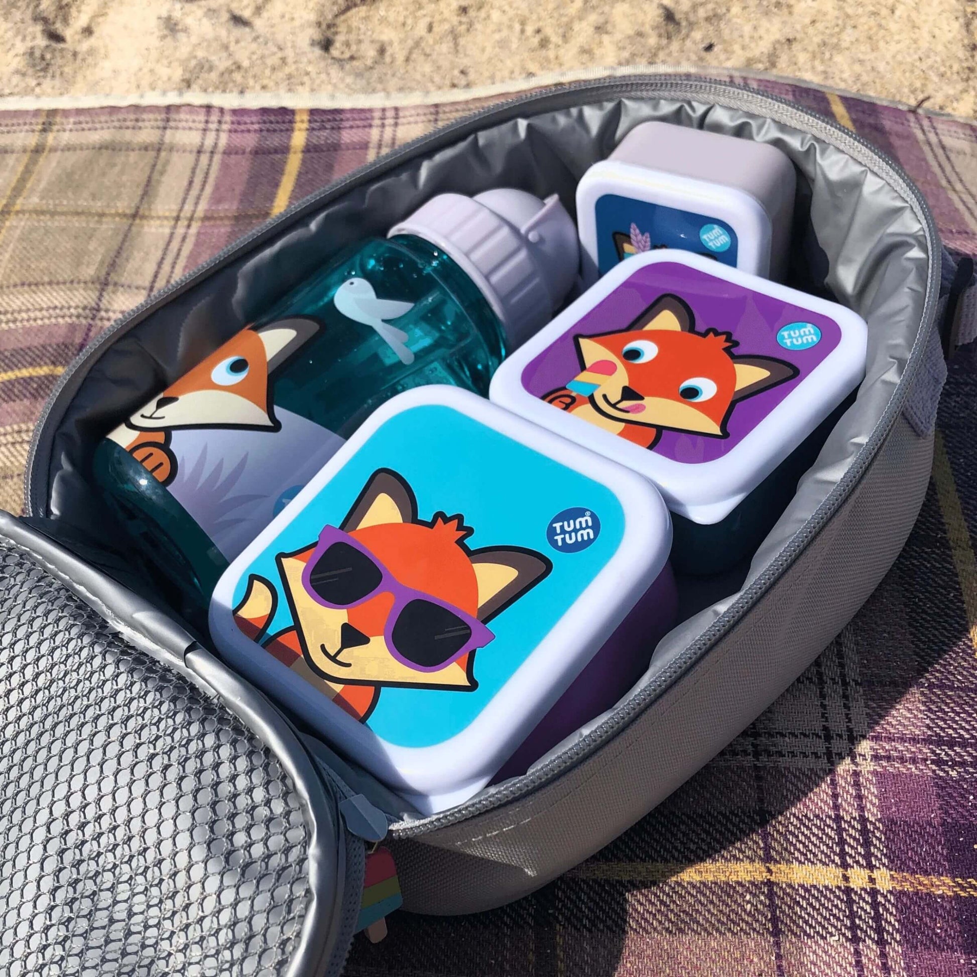 TUM TUM insulated lunch bag, Felicity Fox design, packed with matching snack boxes and water bottle, shown on a plaid blanket for outdoor use.