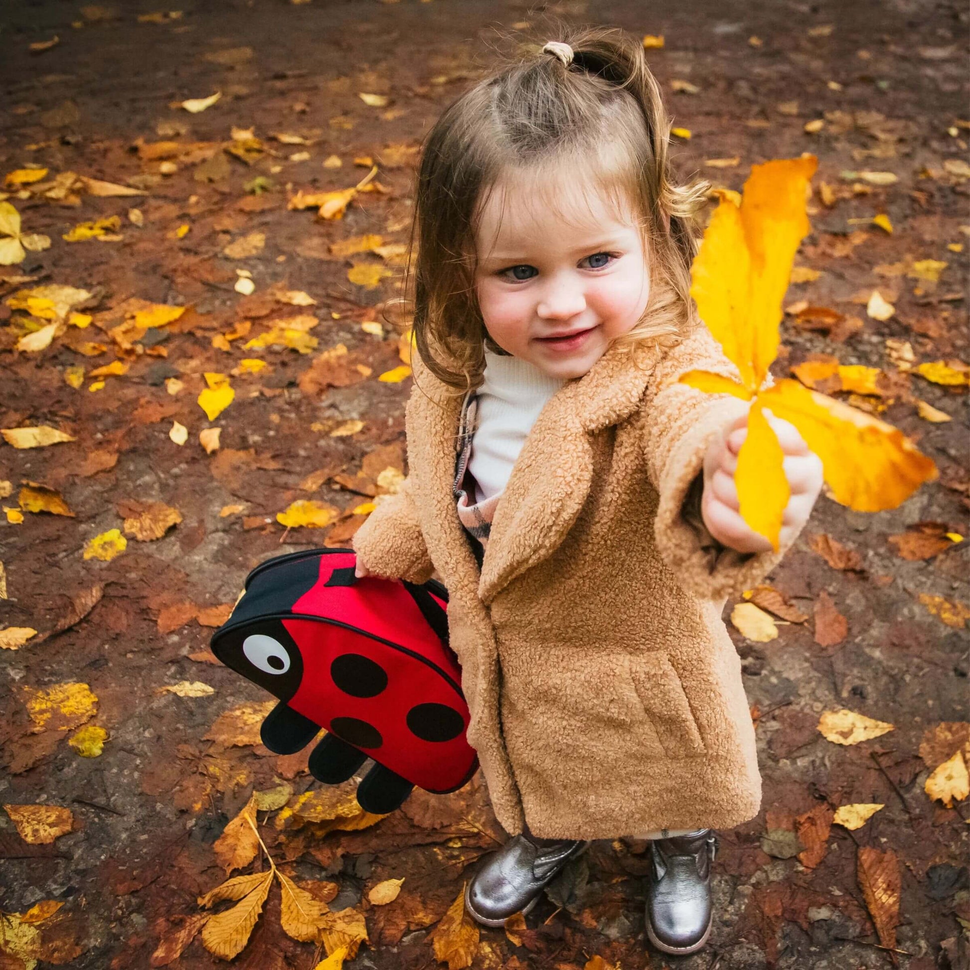 A child holding the TUM TUM Ladybird insulated lunch bag, while exploring autumn leaves outdoors, perfect for nature outings and school lunches.