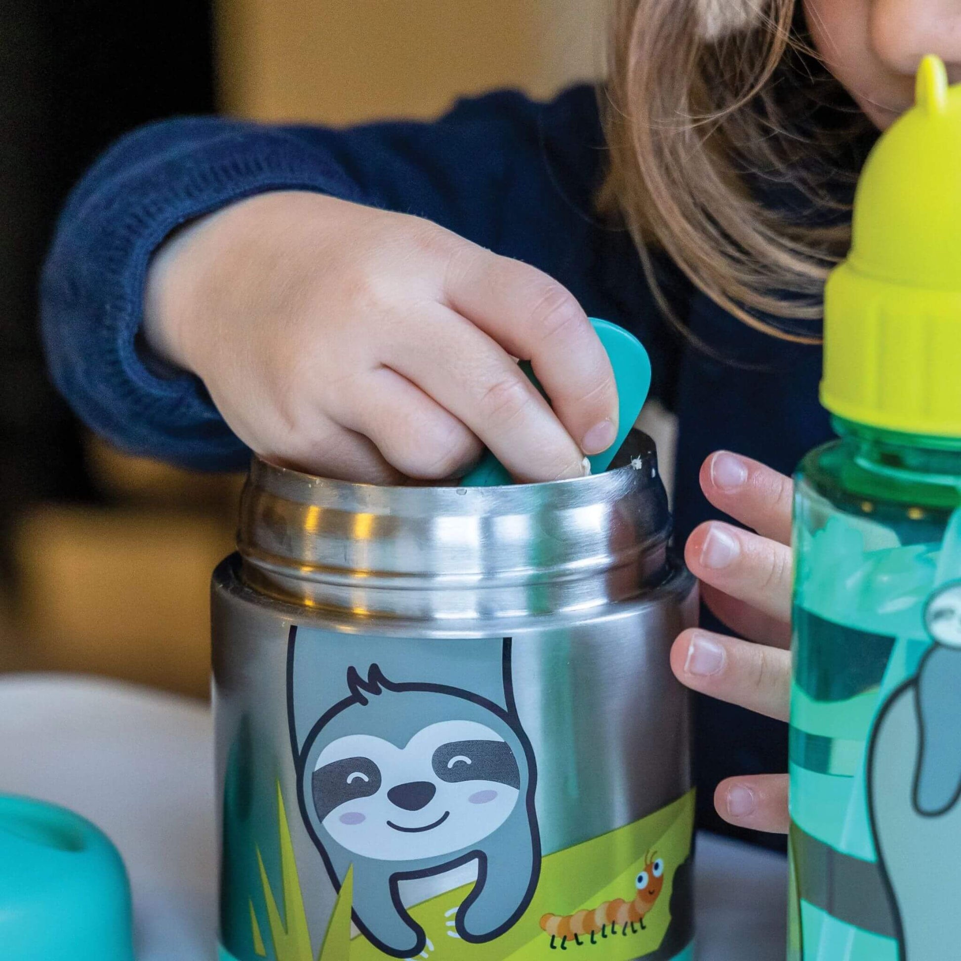 Child using TUM TUM insulated food flask with sloth design, eating from it with a spoon, ideal for keeping kids' food hot or cold.