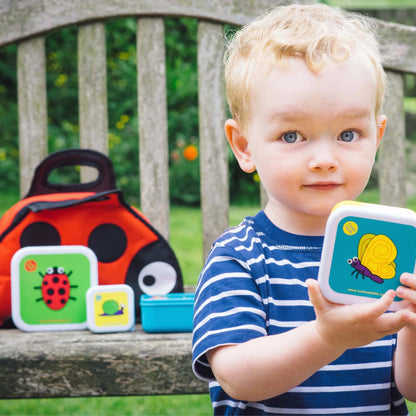 Young child holding TUM TUM Nesting Snack Pots with colourful bug designs, featuring a ladybird lunch bag in the background.