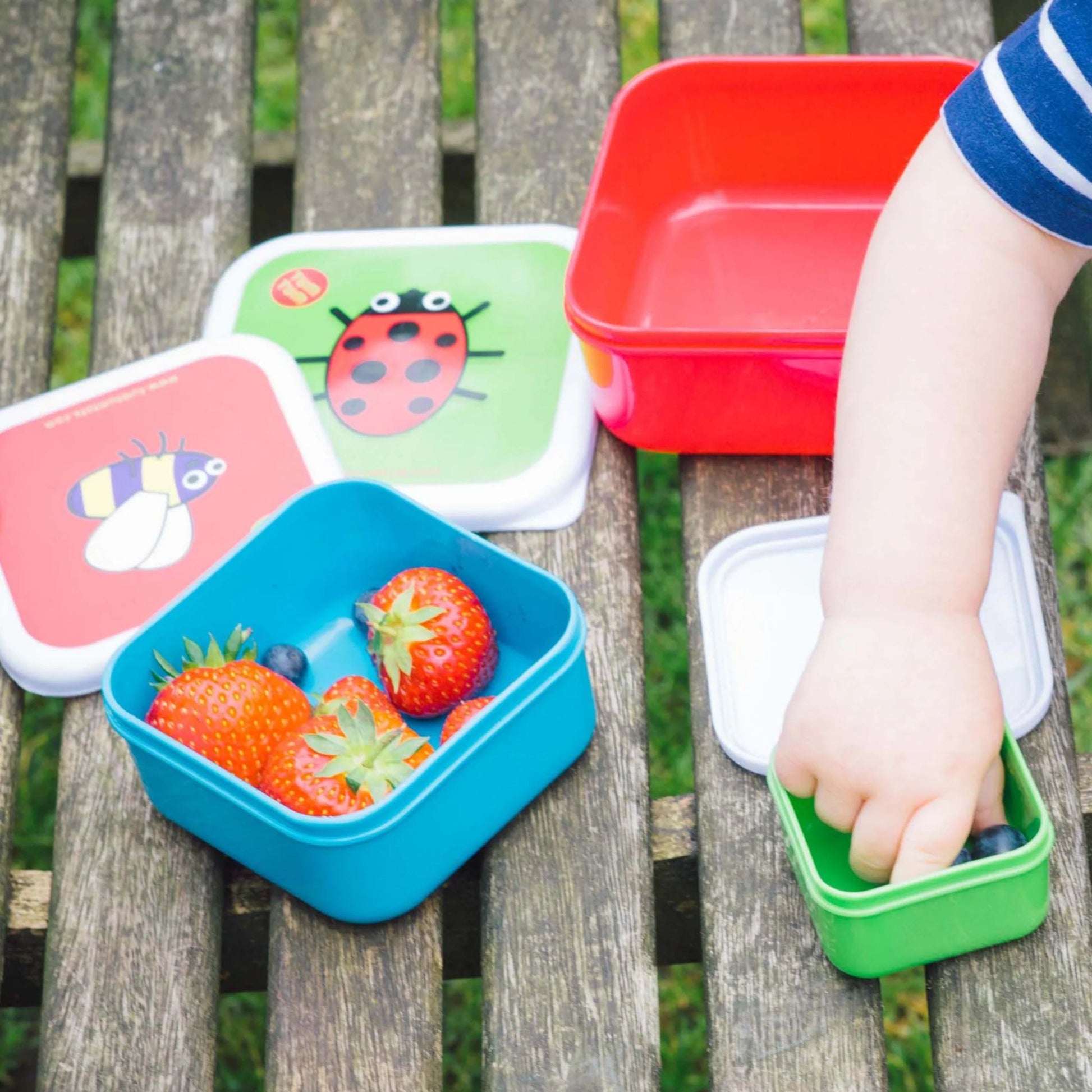 Child reaching into TUM TUM Nesting Snack Pots with colourful bug designs, filled with strawberries and blueberries, ideal for snacks.