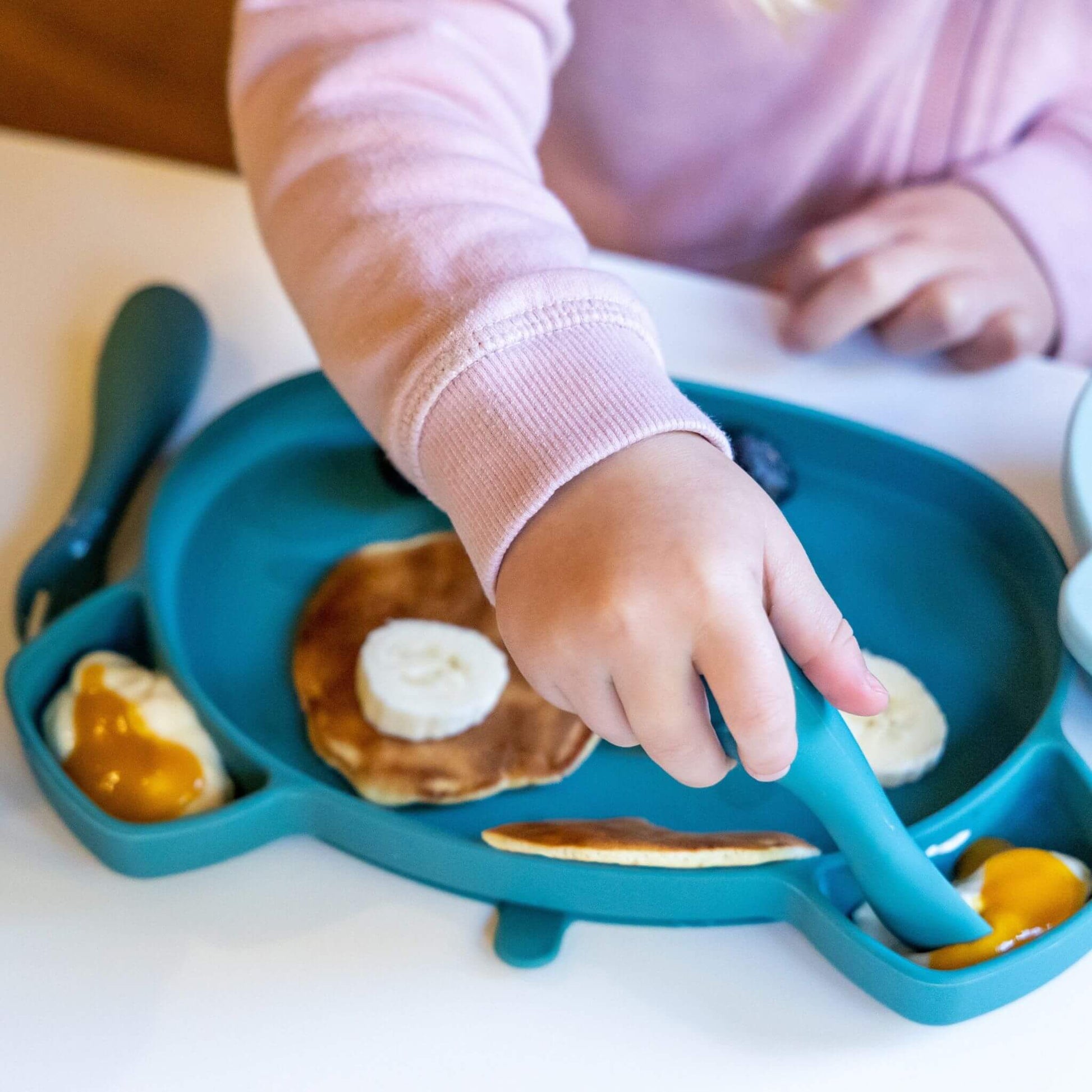 Toddler using blue TUM TUM silicone cutlery and plate to eat pancakes, banana, and yoghurt with fruit, wearing a pink jumper, ideal for self-feeding.