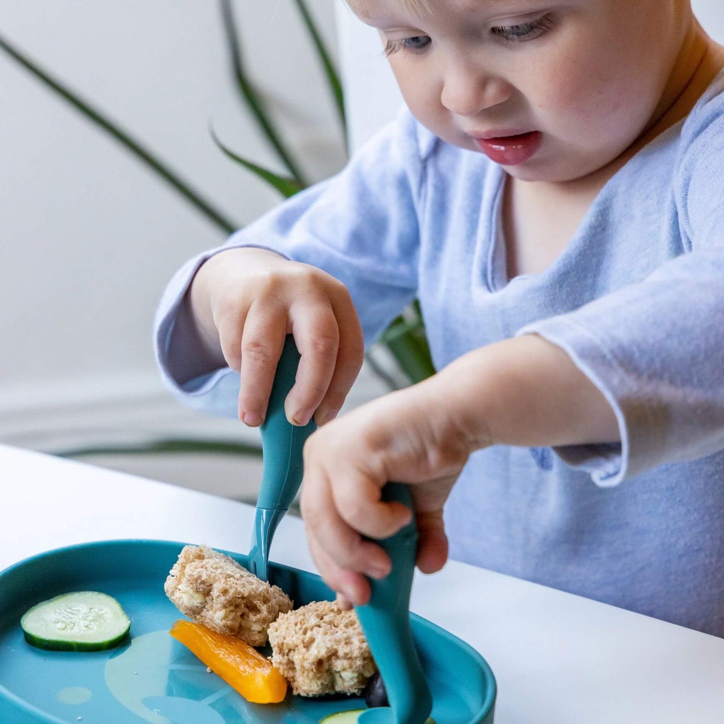 Toddler using blue TUM TUM silicone cutlery with a matching blue plate to eat a sandwich and vegetables, practising self-feeding skills.