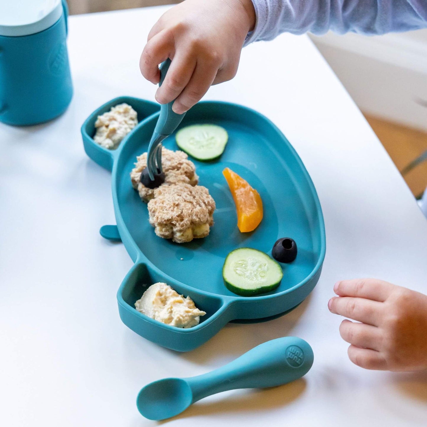 Toddler using blue TUM TUM silicone cutlery and plate with cucumber, sandwich, and hummus, ideal for self-feeding, featuring a matching blue cup.