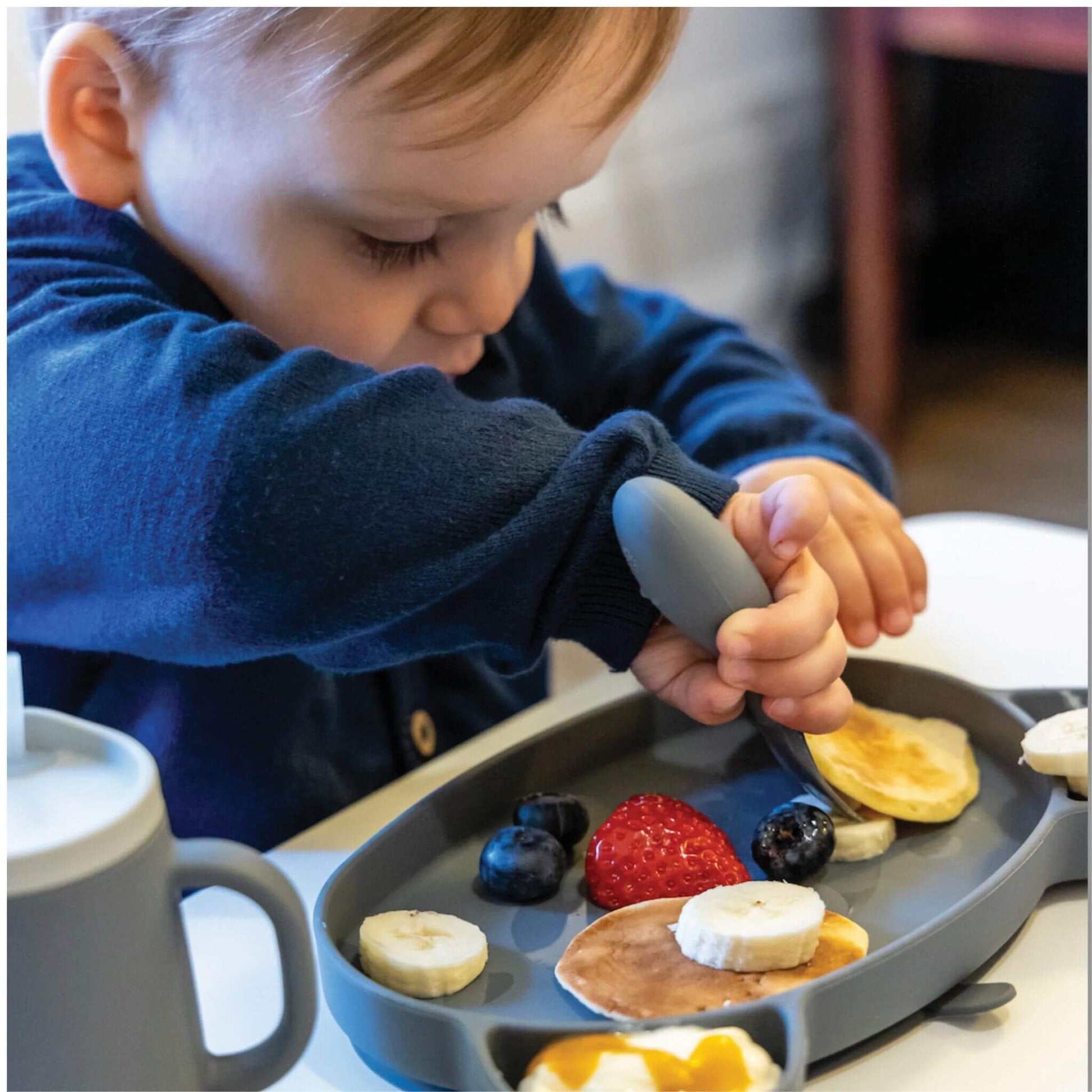 Toddler practising self-feeding with grey TUM TUM silicone cutlery, enjoying pancakes and fruit, promoting independent eating skills.