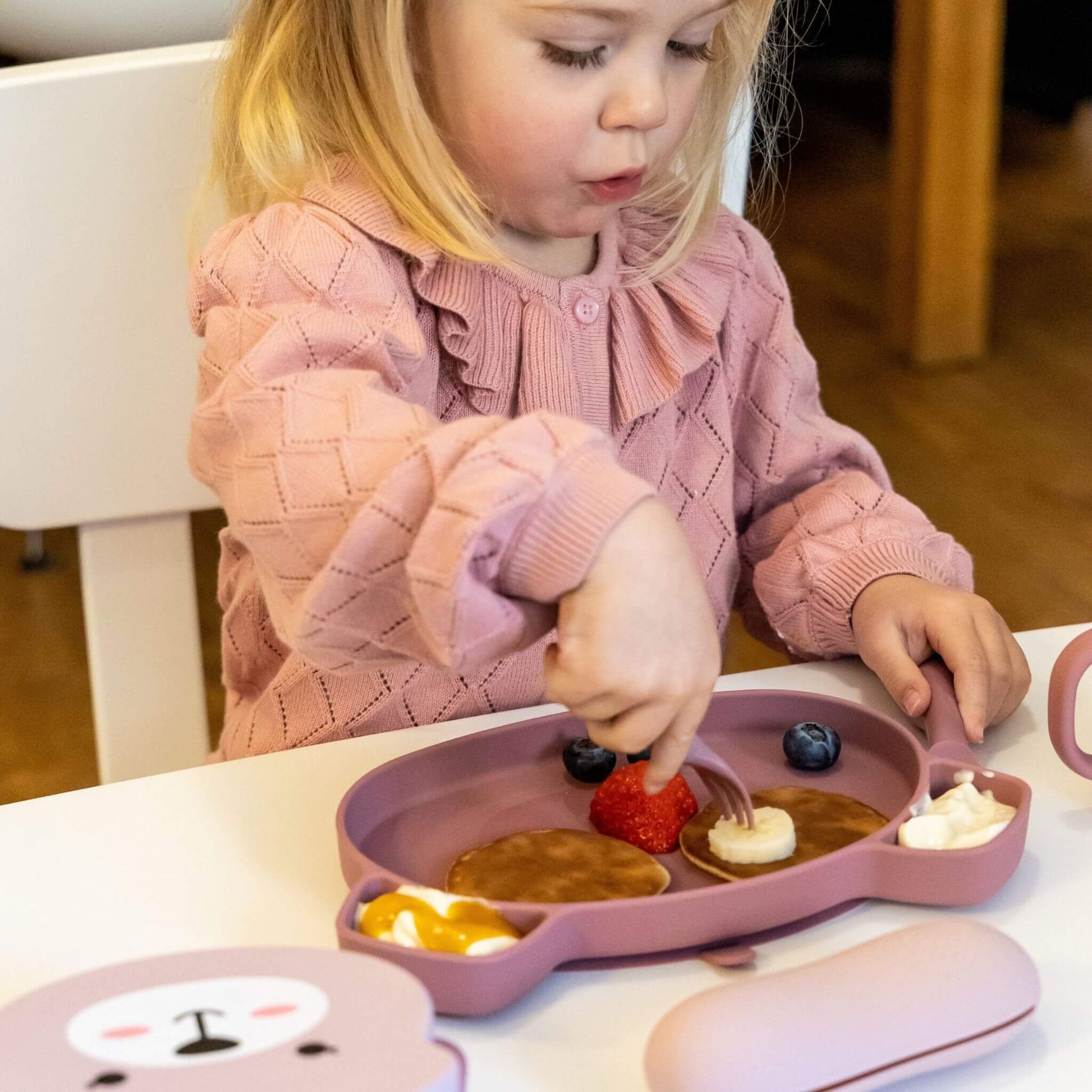 Toddler using pink TUM TUM silicone cutlery and plate with pancakes, banana, and fruit, wearing a pink jumper, ideal for self-feeding.