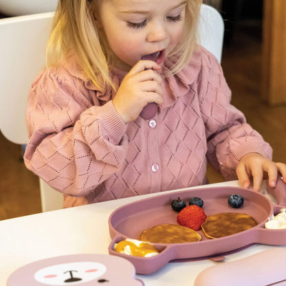 Toddler using pink TUM TUM silicone cutlery and plate with pancakes, fruit, and yoghurt, wearing a pink jumper, perfect for self-feeding.