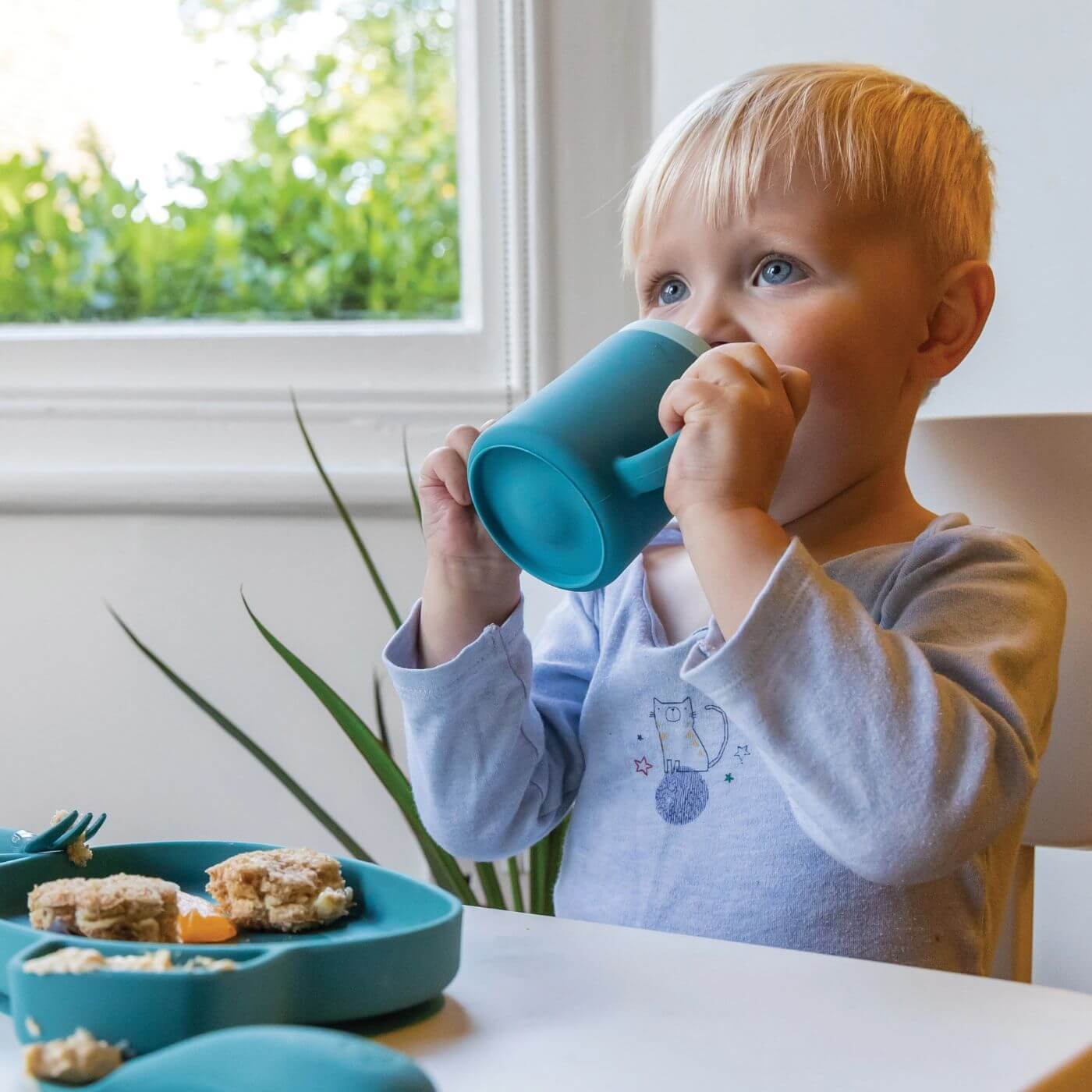 A toddler is drinking from a blue TUM TUM silicone sippy cup with dual handles, designed for easy gripping. The cup is part of a mealtime set, ideal for young children learning to drink independently while enjoying their food.